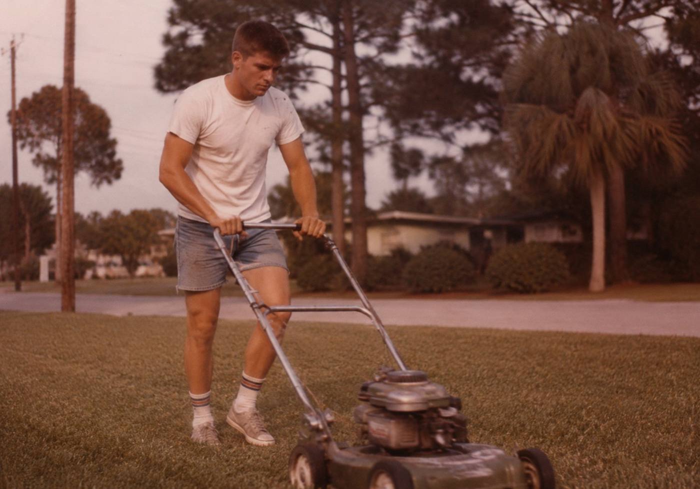 A young Jason mowing a lawn decades ago, white tee and denim shorts, push mower, period photograph