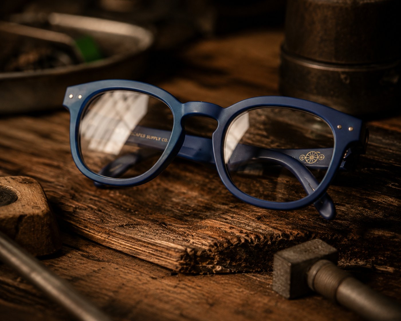 The Scapes eyewear — matte navy acetate frames with a clear lens, SCAPES SUPPLY CO. on the temple and the brushed-gold seedhead glyph, resting on a worn studio workbench.