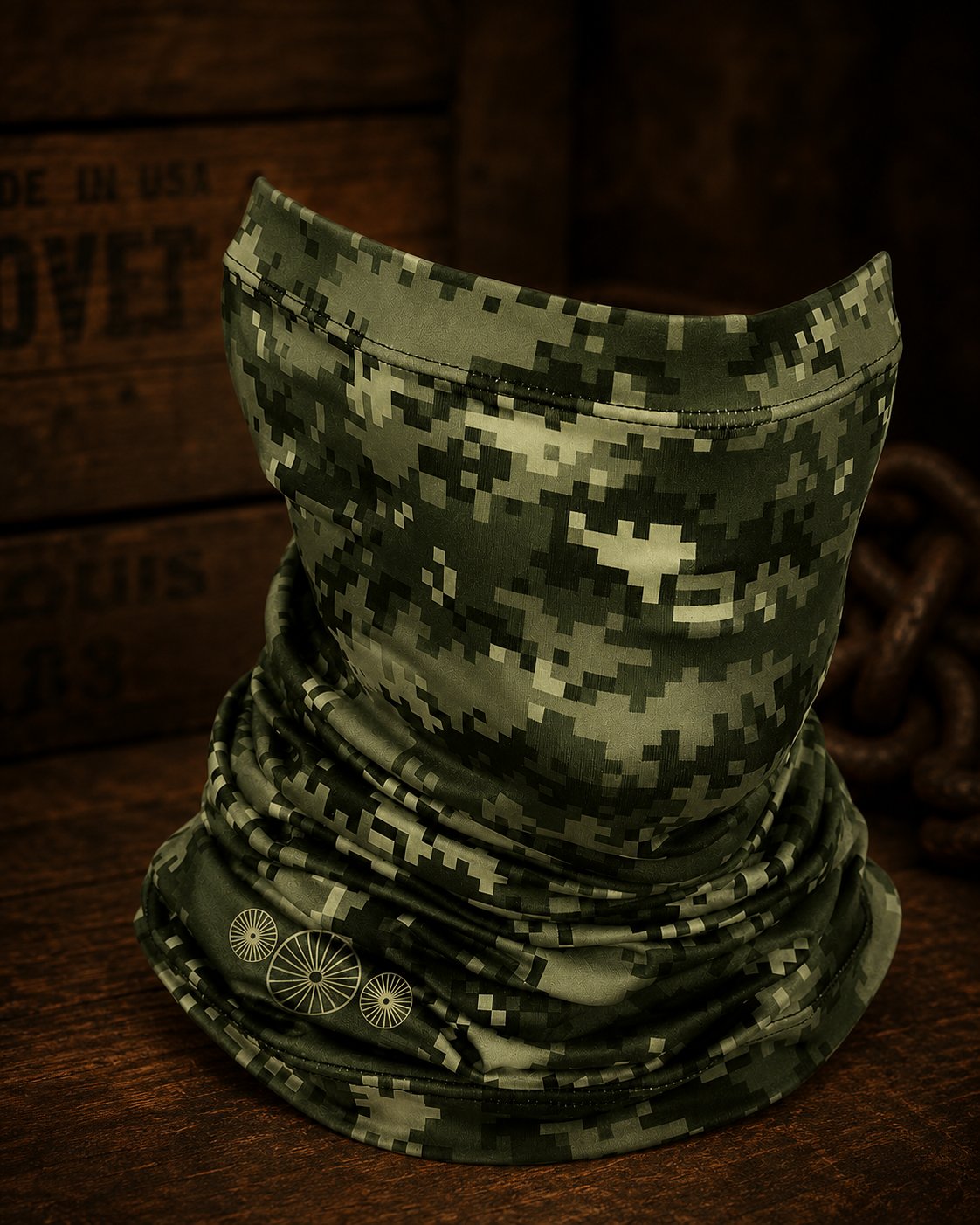The Scapes neck gaiter — digital-camo microfiber printed with the three seedheads at the hem, on a work table beside a chain and a Made-in-USA crate.