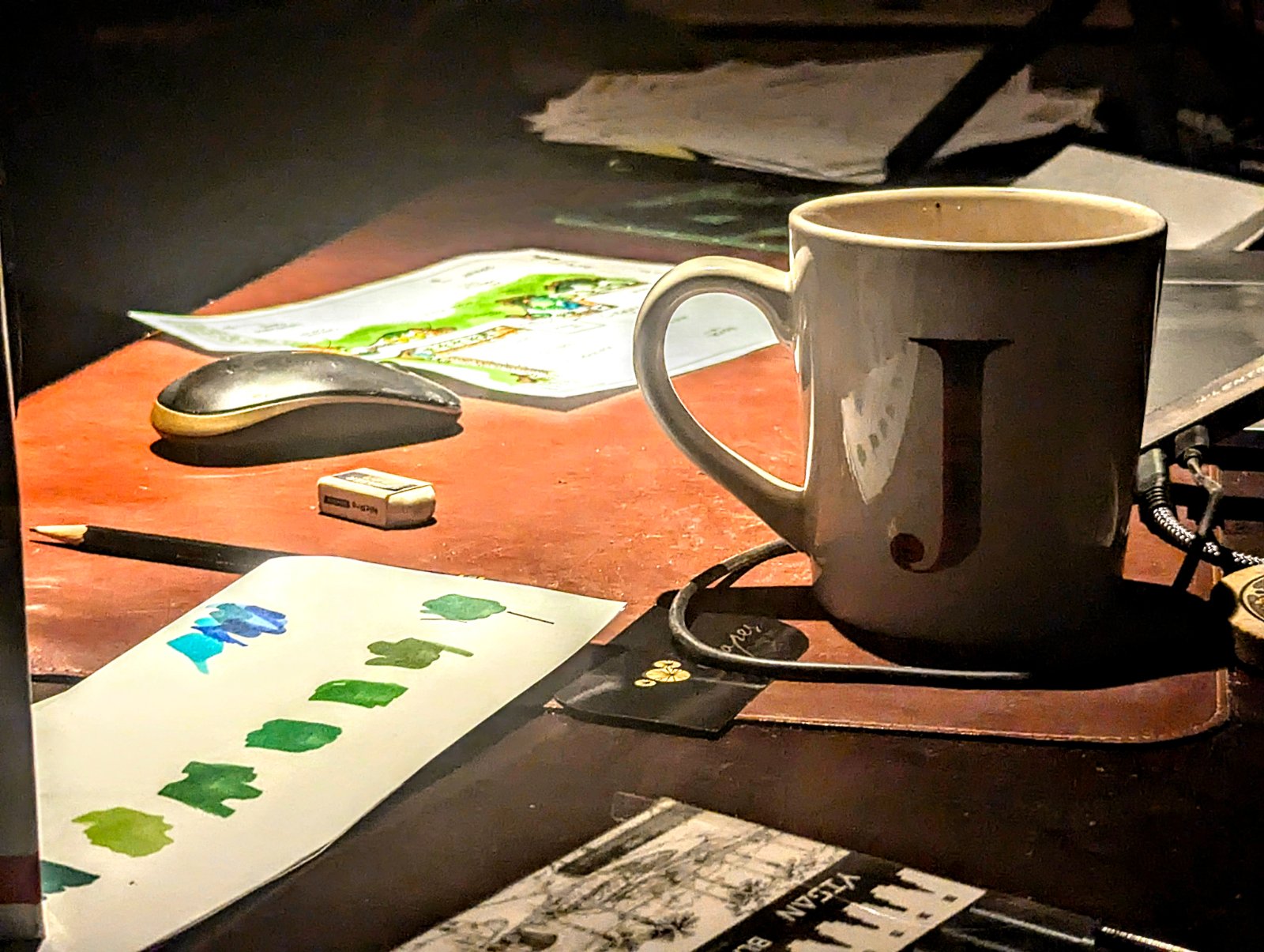 The drafting desk at night — a coffee mug marked with a J, a hand-painted sheet of green and blue watercolor swatches being tested, a landscape concept plan, a pencil and eraser on a worn leather pad